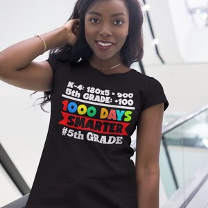 K&ouml;nnte beinhalten: Schwarzes T-Shirt mit dem Text "K-4: 180x5 = 900, 5th GRADE: +100, 1000 DAYS SMARTER #5th GRADE" in Wei&szlig;, Rot, Gelb, Gr&uuml;n und Blau. Das T-Shirt wird von einer Person getragen.