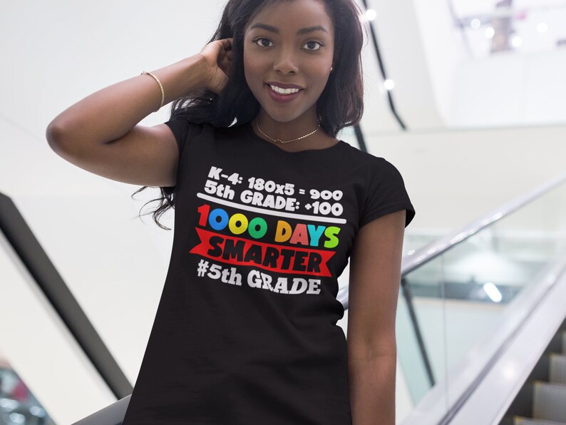 K&ouml;nnte beinhalten: Schwarzes T-Shirt mit dem Text "K-4: 180x5 = 900, 5th GRADE: +100, 1000 DAYS SMARTER #5th GRADE" in Wei&szlig;, Rot, Gelb, Gr&uuml;n und Blau. Das T-Shirt wird von einer Person getragen.