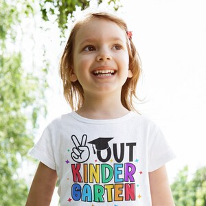 May include: A young girl wearing a white t-shirt with the text "OUT KINDER GARDEN" in rainbow colors and a peace sign. She is smiling and looking up.