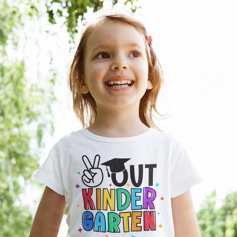 May include: A young girl wearing a white t-shirt with the text "OUT KINDER GARDEN" in rainbow colors and a peace sign. She is smiling and looking up.