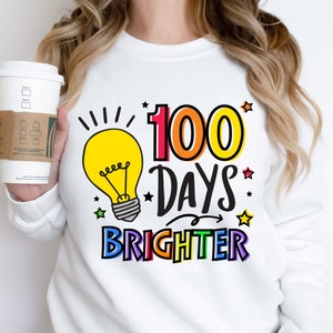 Peut inclure: Sweat-shirt blanc avec un graphique d'ampoule et le texte "100 jours plus lumineux" en couleurs arc-en-ciel.