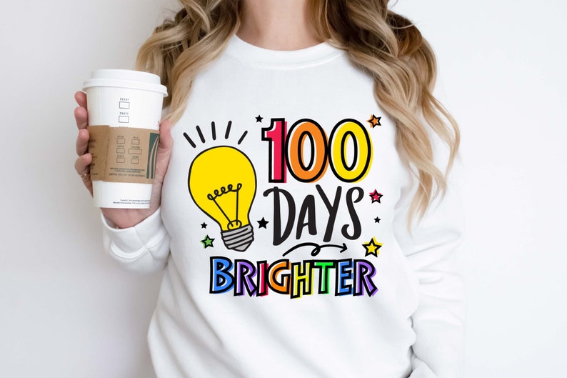 Peut inclure: Sweat-shirt blanc avec un graphique d'ampoule et le texte "100 jours plus lumineux" en couleurs arc-en-ciel.