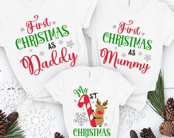 May include: Three white t-shirts with Christmas-themed designs. The first shirt says "First Christmas as Daddy" in red and green text with snowflakes. The second shirt says "First Christmas as Mummy" in red and green text with snowflakes. The third shirt says "My 1st Christmas" in green text with a red and white candy cane and a reindeer.