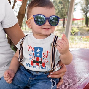 May include: A white t-shirt with a red, white, and blue American flag design. The shirt says "My 1st 4th of July" with a black bow tie on the flag. A young child is wearing the shirt and sunglasses.