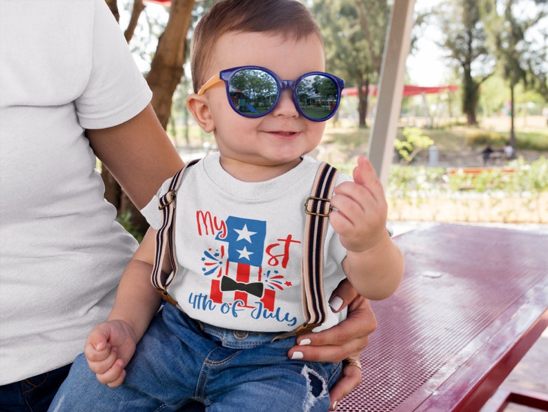 May include: A white t-shirt with a red, white, and blue American flag design. The shirt says "My 1st 4th of July" with a black bow tie on the flag. A young child is wearing the shirt and sunglasses.
