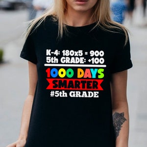 K&ouml;nnte beinhalten: Schwarzes T-Shirt mit dem Text "K-4: 180x5 = 900, 5th GRADE: +100, 1000 DAYS SMARTER, #5th GRADE" in Wei&szlig;, Rot, Blau und Gr&uuml;n. Das T-Shirt hat einen Rundhalsausschnitt.