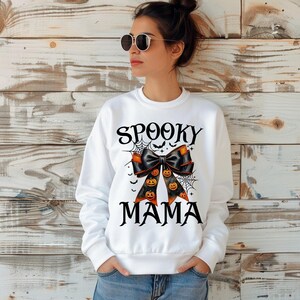 May include: White sweatshirt with the text "Spooky Mama" and a black bow with orange pumpkins on it.