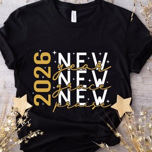 Peut inclure: T-shirt noir avec texte dor&eacute; et blanc. Le motif comprend l'ann&eacute;e "2026" et les mots "NEW year NEW grace NEW praise". Le t-shirt a un col rond et des manches courtes. Des &eacute;toiles dor&eacute;es d&eacute;coratives sont au premier plan.