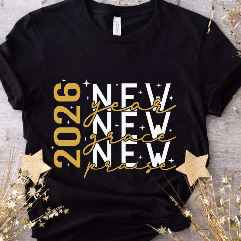 Peut inclure: T-shirt noir avec texte dor&eacute; et blanc. Le motif comprend l'ann&eacute;e "2026" et les mots "NEW year NEW grace NEW praise". Le t-shirt a un col rond et des manches courtes. Des &eacute;toiles dor&eacute;es d&eacute;coratives sont au premier plan.