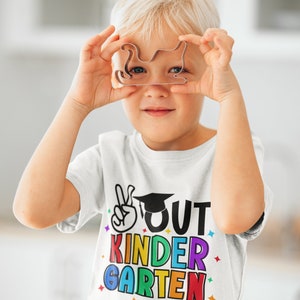 May include: A young child wearing a white t-shirt with the text "OUT KINDER GARDEN" in rainbow colors and a graduation cap graphic. The child is holding a metal cookie cutter in front of their face.
