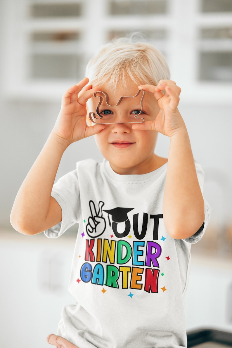 May include: A young child wearing a white t-shirt with the text "OUT KINDER GARDEN" in rainbow colors and a graduation cap graphic. The child is holding a metal cookie cutter in front of their face.