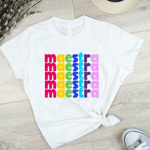 May include: White t-shirt with the word "maestra" repeated five times in rainbow colours.
