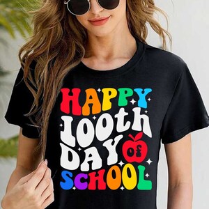 以下が含まれることがあります： 「Happy 100th Day of School」と赤いリンゴのグラフィックが描かれたレインボーカラーのグラフィックが施された黒いTシャツです。