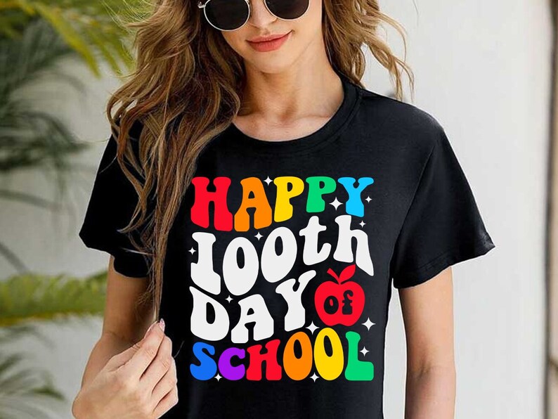 以下が含まれることがあります： 「Happy 100th Day of School」と赤いリンゴのグラフィックが描かれたレインボーカラーのグラフィックが施された黒いTシャツです。
