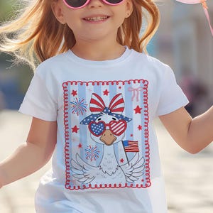 May include: White t-shirt with a red, white, and blue goose wearing heart-shaped sunglasses and a bow. The goose is holding an American flag. The design is surrounded by a red, white, and blue border.
