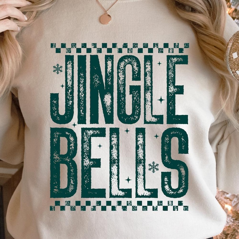 May include: Cream-colored sweatshirt with the text "JINGLE BELLS" in distressed teal lettering. The design includes snowflakes and a checkered border, creating a festive holiday theme. The sweatshirt is a comfortable, casual garment.