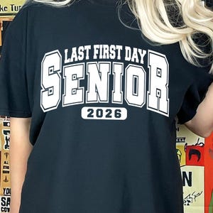 May include: Black t-shirt with white text reading "LAST FIRST DAY SENIOR 2026". The text is in a collegiate style font. The shirt is a short-sleeved crew neck.