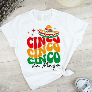May include: White t-shirt featuring a colourful graphic design that reads "Cinco Cinco Cinco de Mayo" with a sombrero on top.