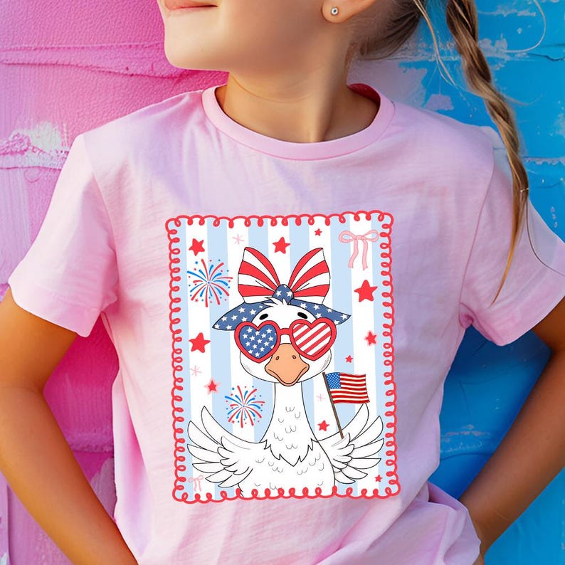 May include: A pink t-shirt with a white goose wearing red, white, and blue heart-shaped sunglasses and a bow. The goose is holding a small American flag. The design is surrounded by a red, white, and blue border with stars and stripes.