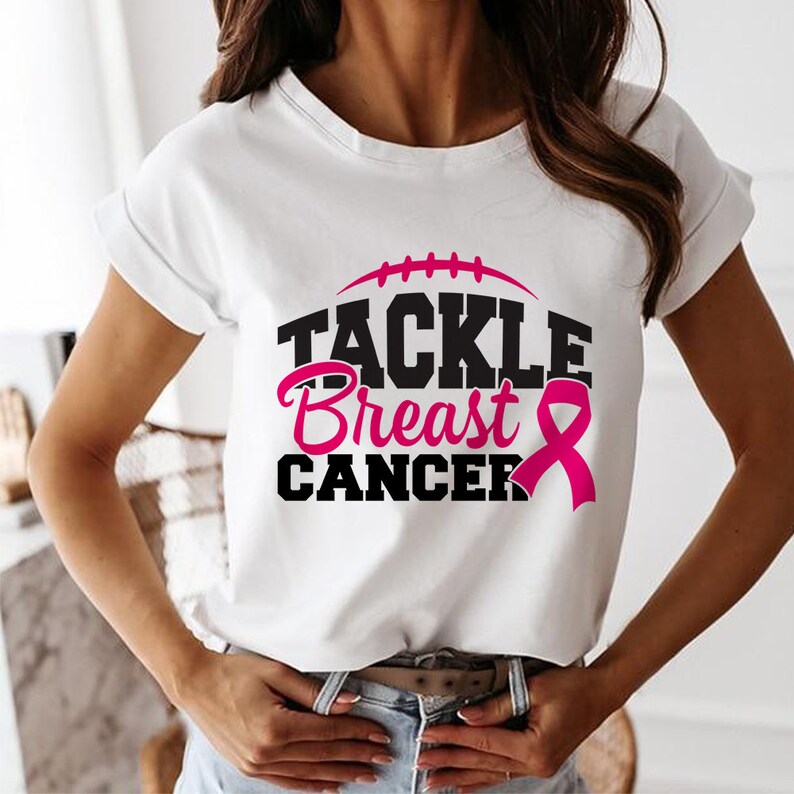 Peut inclure: T-shirt blanc avec un motif noir qui dit "Tackle Breast Cancer" avec un ruban rose. Le motif comprend &eacute;galement un ballon de football noir.