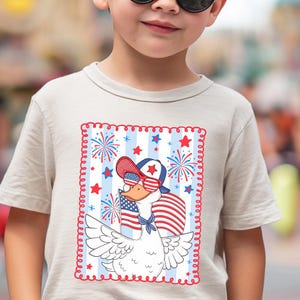 May include: A light gray t-shirt with a cartoon goose wearing sunglasses and a red, white, and blue baseball cap. The goose is holding an American flag and is surrounded by fireworks and stars. The design is framed with a red and blue stitched border.