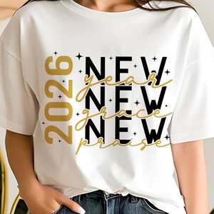 Peut inclure: T-shirt blanc avec l'inscription "NEW year grace NEW praise" en noir et or. L'ann&eacute;e "2026" est imprim&eacute;e verticalement en or sur le c&ocirc;t&eacute; gauche. De petites &eacute;toiles ornent le texte.
