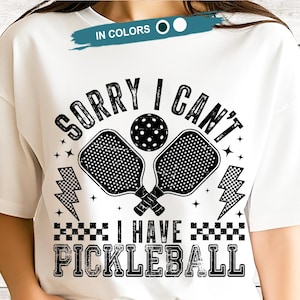 May include: White t-shirt with a black graphic design featuring pickleball paddles, a ball, and the text "SORRY I CAN'T I HAVE PICKLEBALL". The design includes lightning bolts and stars.
