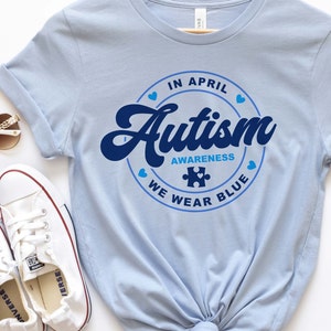 Puede incluir: Camiseta azul claro con un gr&aacute;fico circular azul que dice "En abril, conciencia del autismo, usamos azul" con una pieza de rompecabezas azul en el centro.