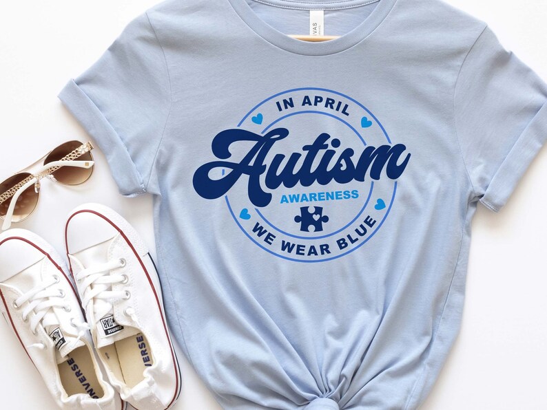 Puede incluir: Camiseta azul claro con un gr&aacute;fico circular azul que dice "En abril, conciencia del autismo, usamos azul" con una pieza de rompecabezas azul en el centro.
