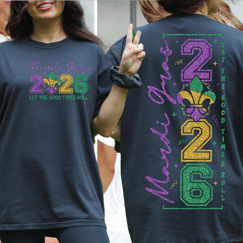 Op de afbeelding: Donker marineblauw T-shirt met een Mardi Gras thema. De voorkant heeft de tekst "Mardi Gras 2026" met een fleur-de-lis ontwerp in paars, groen en goud. De achterkant toont "Mardi Gras" verticaal en "2026" met een fleur-de-lis.
