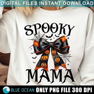 May include: A white sweatshirt with the text "Spooky Mama" in black. A black and orange bow with a spiderweb design and orange pumpkins is centered on the shirt.