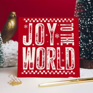 May include: A red square Christmas card with the words "JOY TO THE WORLD" in white, distressed font. The card has a decorative border and snowflake accents. A gold pen and paper clip are in the foreground.
