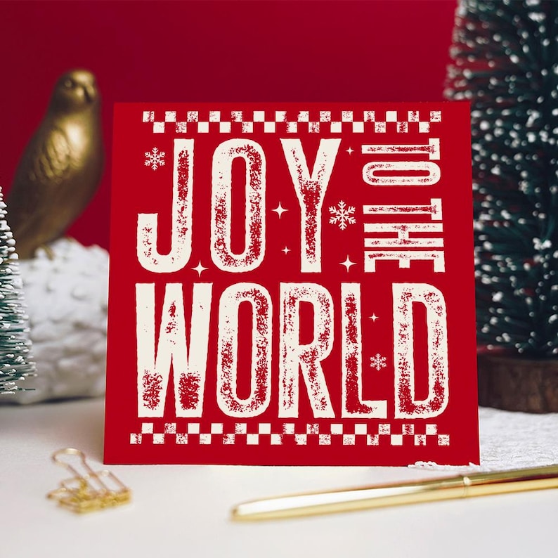 May include: A red square Christmas card with the words "JOY TO THE WORLD" in white, distressed font. The card has a decorative border and snowflake accents. A gold pen and paper clip are in the foreground.