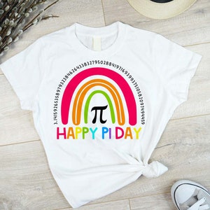 May include: White t-shirt with a rainbow design and the pi symbol in the centre. The words "HAPPY PI DAY" are printed below the rainbow. Numbers representing pi's value are arched above the rainbow. A fun, colourful design.