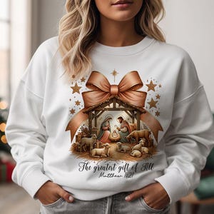 May include: White sweatshirt with a brown bow and a nativity scene graphic. The graphic includes a manger, Mary, Joseph, baby Jesus, and sheep. The text "The greatest gift of all Matthew 1:21" is printed below the graphic.