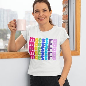 May include: A white t-shirt with the word "maestra" repeated five times in rainbow colours. The shirt is being worn by a person holding a pink mug.