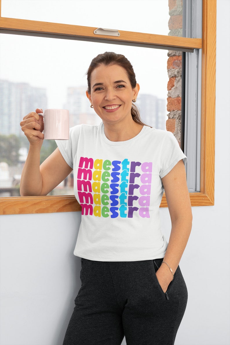 May include: A white t-shirt with the word "maestra" repeated five times in rainbow colours. The shirt is being worn by a person holding a pink mug.