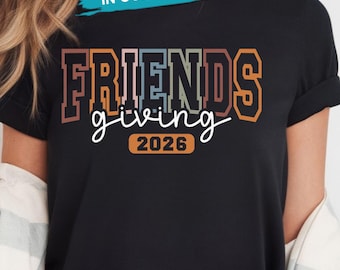 May include: A black short-sleeved t-shirt featuring the text FRIENDS in a multi-colored block font, the word giving in white cursive script, and the year 2026 inside an orange rounded rectangle. The design uses muted tones of pink, blue, and brown.