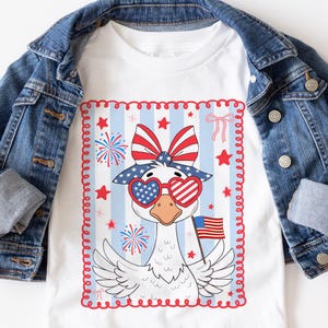 May include: A white t-shirt with a red, white, and blue goose wearing heart-shaped sunglasses and a bow. The goose is holding a small American flag. The design is surrounded by a red, white, and blue border.