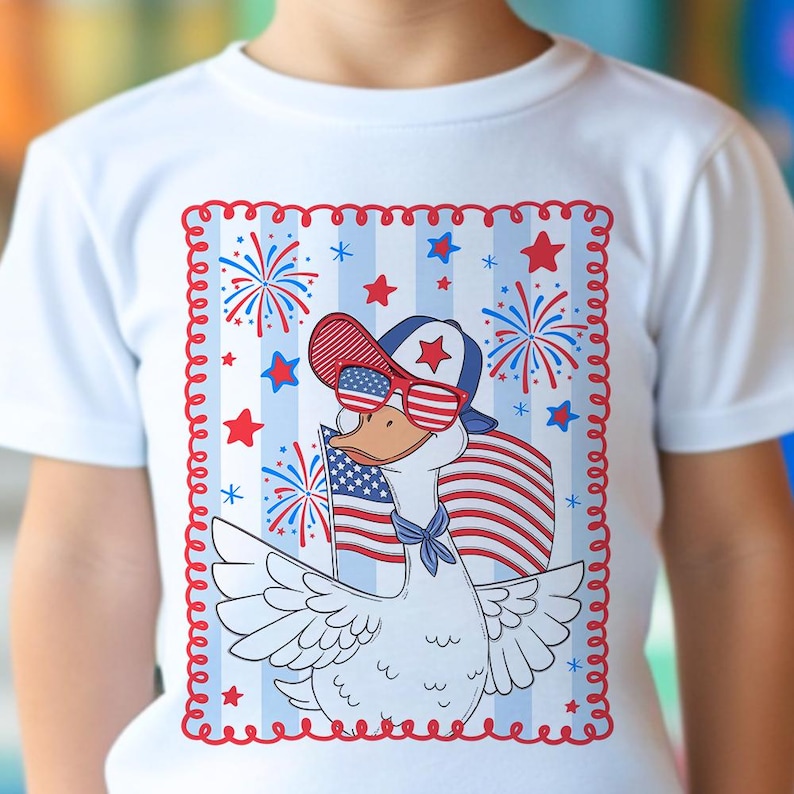 May include: A white t-shirt with a red, white, and blue illustration of a goose wearing sunglasses and a baseball cap. The goose is holding an American flag and there are fireworks in the background.
