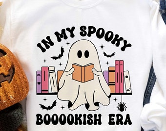 Könnte beinhalten: Ein weißes Sweatshirt mit einem Geist, der ein Buch liest, umgeben von Bücherregalen. Der Text lautet "In My Spooky Booookish Era".