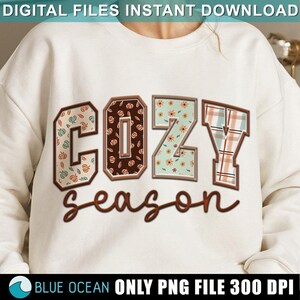 May include: A white sweatshirt with the text "Cozy season" in brown lettering. The letters are decorated with fall-themed patterns, including pumpkins, flowers, and plaid.