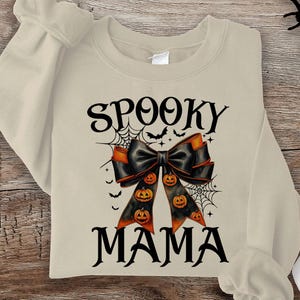 May include: A beige sweatshirt with the text "Spooky Mama" in black. A black bow with orange accents and pumpkins is centered on the sweatshirt.