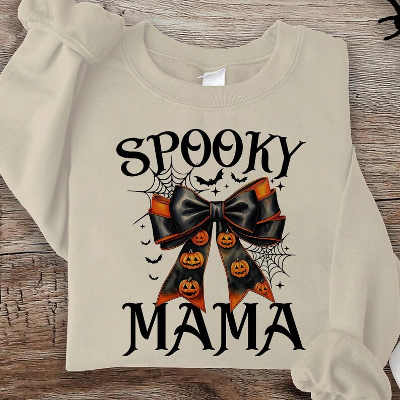 May include: A beige sweatshirt with the text "Spooky Mama" in black. A black bow with orange accents and pumpkins is centered on the sweatshirt.