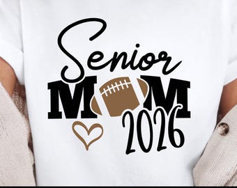 Pode incluir: Camiseta branca com um gráfico preto e castanho. O design inclui as palavras "Senior Mom" com uma bola de futebol no lugar do "O" em Mom, um coração e o ano "2026". A parte inferior da camiseta tem as palavras "BLUE OCEAN" e "SVG PNG DXF JPG EPS".