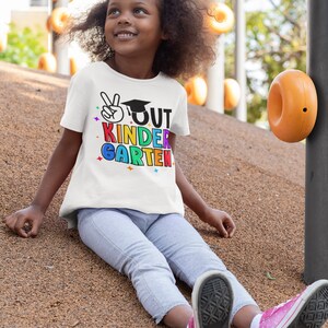 May include: A young girl wearing a white t-shirt with the text "OUT KINDER-GARTEN" in rainbow colors and a peace sign with a graduation cap. She is sitting on a playground surface with orange playground equipment in the background.