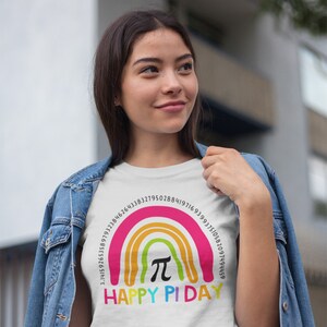 May include: White t-shirt featuring a rainbow design with the text "HAPPY PI DAY." The rainbow includes the mathematical symbol &pi; and a series of numbers. The shirt is worn by a person wearing a denim jacket.
