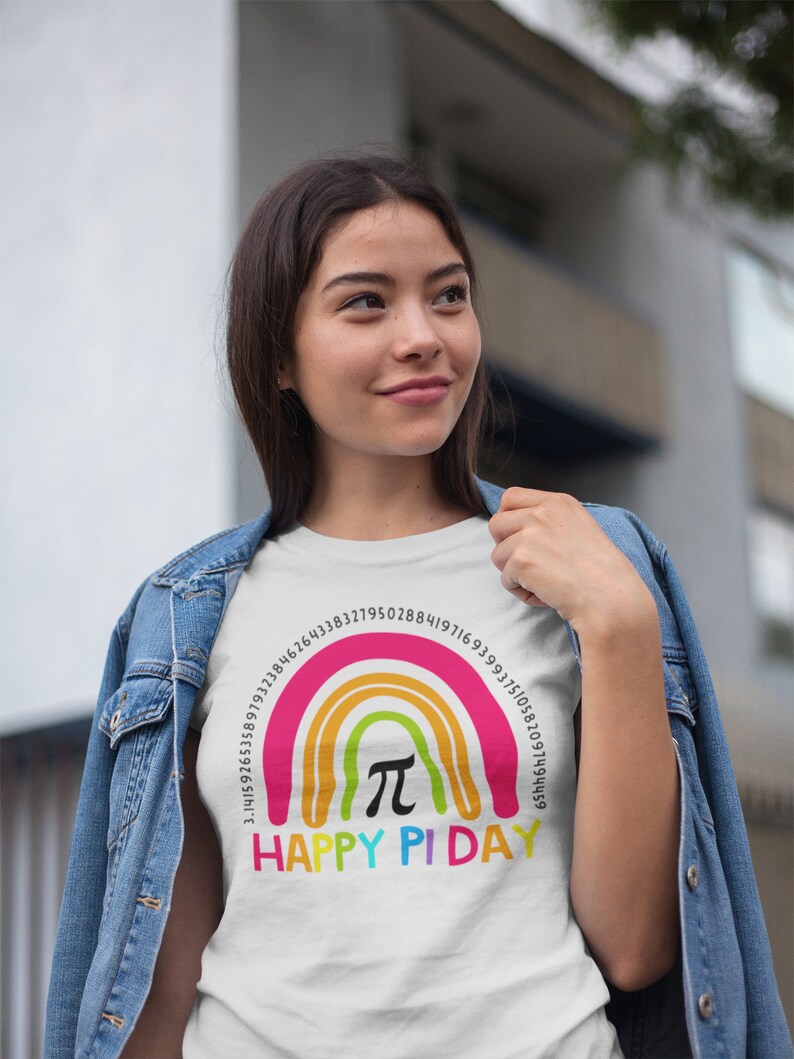 May include: White t-shirt featuring a rainbow design with the text "HAPPY PI DAY." The rainbow includes the mathematical symbol &pi; and a series of numbers. The shirt is worn by a person wearing a denim jacket.