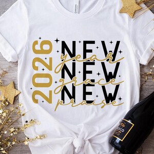 Peut inclure: T-shirt blanc avec l'inscription "2026 New Year New Grace New Praise" en or et noir. Le t-shirt a un col rond et des manches courtes. Le motif comprend des &eacute;toiles.