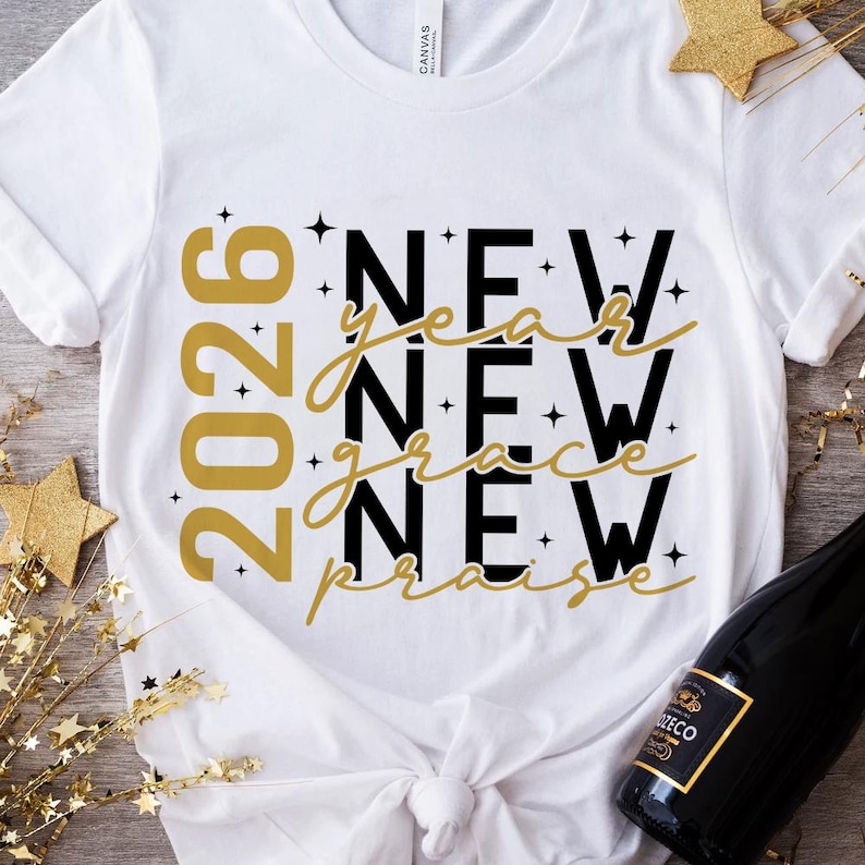 Peut inclure: T-shirt blanc avec l'inscription "2026 New Year New Grace New Praise" en or et noir. Le t-shirt a un col rond et des manches courtes. Le motif comprend des &eacute;toiles.